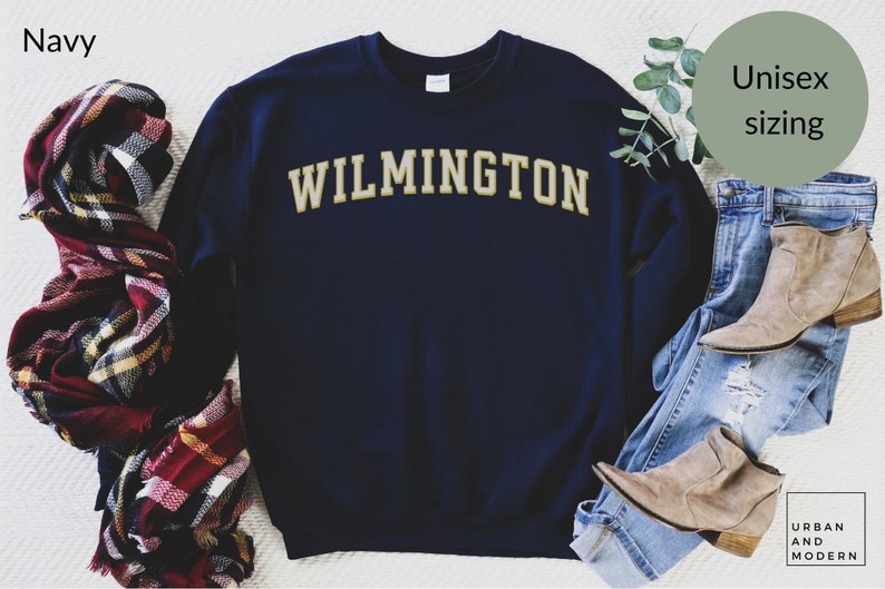 May include: Navy blue sweatshirt with "WILMINGTON" in gold lettering. The image includes a plaid scarf, jeans, and boots. The sweatshirt is labeled "Unisex sizing". The brand name "URBAN AND MODERN" is also visible.