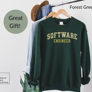 May include: A forest green sweatshirt with the words "SOFTWARE ENGINEER" in gold lettering. The sweatshirt is displayed on a wooden hanger alongside other garments. A green circle with the text "Great Gift!" is visible.