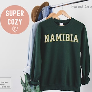 May include: Forest green sweatshirt with the word "NAMIBIA" in gold lettering. The sweatshirt is hanging on a rack with other clothing.