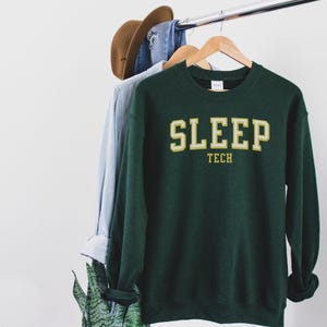 May include: A dark green sweatshirt with the text "SLEEP TECH" in a gold and white collegiate-style font. The sweatshirt is hanging on a wooden hanger with other clothing items.