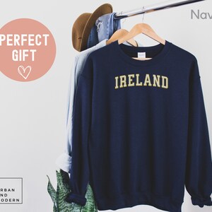 Może przedstawiać: Granatowa bluza z napisem "IRELAND" w złotych literach. Bluza wisi na drewnianym wieszaku z innymi ubraniami. Tekst "PERFECT GIFT" znajduje się w r&oacute;żowym okręgu.