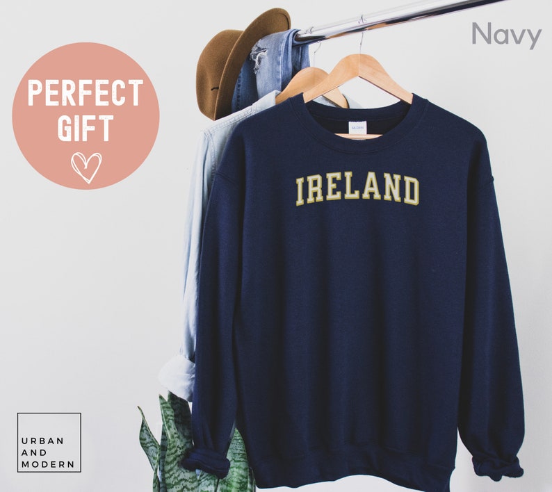Może przedstawiać: Granatowa bluza z napisem "IRELAND" w złotych literach. Bluza wisi na drewnianym wieszaku z innymi ubraniami. Tekst "PERFECT GIFT" znajduje się w r&oacute;żowym okręgu.