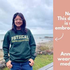 Op de afbeelding: Een groen sweatshirt met de tekst "PHYSICAL MEDICINE & REHABILITATION" in gele letters. Het model draagt maat M.