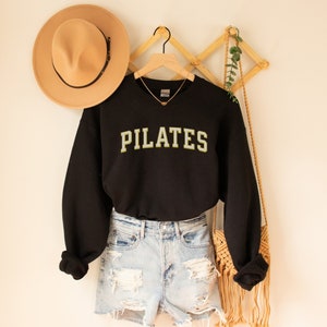 May include: Black sweatshirt with the word "PILATES" in yellow and white letters. The sweatshirt is hanging on a wooden hanger and is paired with denim shorts.