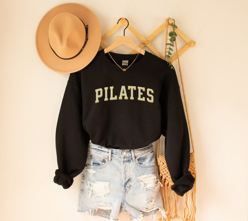 May include: Black sweatshirt with the word "PILATES" in yellow and white letters. The sweatshirt is hanging on a wooden hanger and is paired with denim shorts.