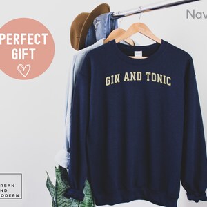May include: Navy blue sweatshirt with the text "GIN AND TONIC" in gold lettering. The sweatshirt is hanging on a wooden hanger. A light blue shirt and a brown hat are also hanging on the rack. The words "PERFECT GIFT" are in a pink circle.