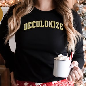 May include: Black long-sleeve shirt with the word "DECOLONIZE" in gold lettering. The person is holding a white enamel mug with marshmallows and a candy cane. The image has a "Great Gift" sticker.