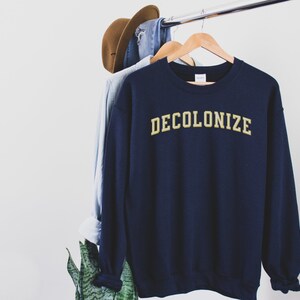 Pu&ograve; includere: Una felpa blu navy con la parola "DECOLONIZE" stampata in lettere dorate sul davanti.