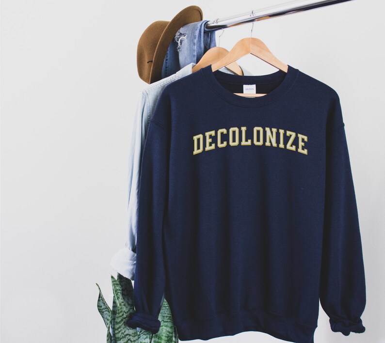 Pu&ograve; includere: Una felpa blu navy con la parola "DECOLONIZE" stampata in lettere dorate sul davanti.
