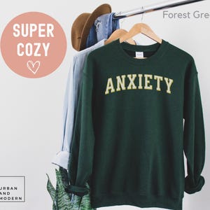 May include: Forest green sweatshirt with the word "ANXIETY" in gold lettering. The sweatshirt is hanging on a wooden hanger with a light blue shirt and a brown hat. The words "SUPER COZY" are in a pink circle.