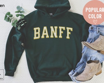 May include: A dark green hooded sweatshirt with the word "BANFF" in yellow letters with a white outline. The sweatshirt is laid flat on a white surface with a pair of blue jeans and brown ankle boots.