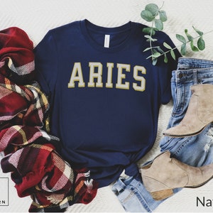 Op de afbeelding: Marineblauw t-shirt met het woord "ARIES" in gouden letters. Het shirt wordt getoond met een rood, bordeauxrood en geel geruite sjaal, een spijkerbroek en beige enkellaarsjes. De tekst "Urban and Modern" staat linksonder.