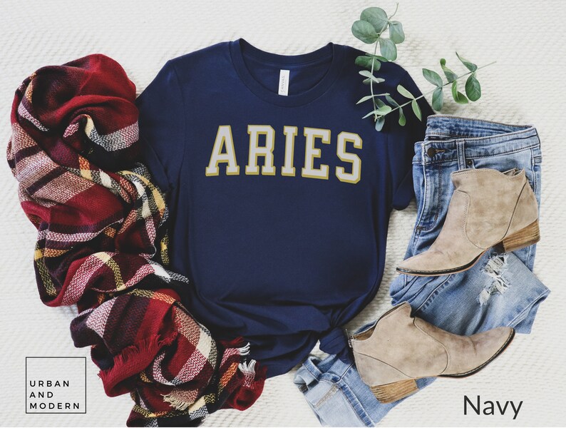 Op de afbeelding: Marineblauw t-shirt met het woord "ARIES" in gouden letters. Het shirt wordt getoond met een rood, bordeauxrood en geel geruite sjaal, een spijkerbroek en beige enkellaarsjes. De tekst "Urban and Modern" staat linksonder.