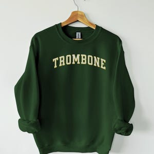 May include: A dark green sweatshirt with the word "TROMBONE" printed in a gold, arched font. The sweatshirt is hanging on a wooden hanger against a white background. The sleeves are cuffed.