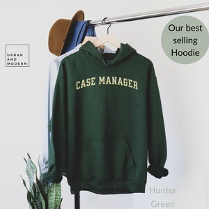May include: Hunter green hoodie with the text "CASE MANAGER" in a gold arched font. The hoodie has a front pocket and a hood. The image also includes a brown hat and a denim shirt hanging on a clothing rack. The text "Our best selling Hoodie" is also visible.