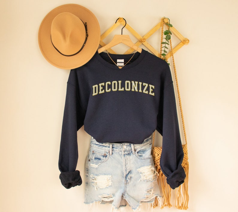 Pu&ograve; includere: Una felpa blu navy con la scritta "DECOLONIZE" in caratteri dorati. La felpa &egrave; indossata sopra un paio di pantaloncini di jeans blu chiaro.