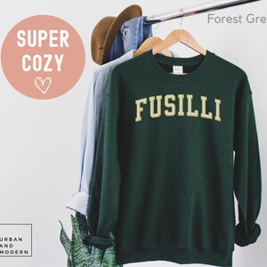 May include: A forest green sweatshirt with the word "FUSILLI" in a gold, collegiate-style font. The sweatshirt is hanging on a wooden hanger with other clothing. The words "SUPER COZY" are in a pink circle with a heart.