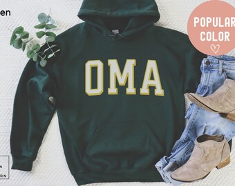 May include: A dark green hoodie with the word "OMA" in a white and yellow block letter design. The hoodie is displayed with blue jeans and tan boots. The words "Popular Color" are in a pink circle.