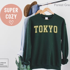 May include: Forest green sweatshirt with the word "TOKYO" in a gold and white collegiate font. The sweatshirt is hanging on a wooden hanger with other clothing items. The words "SUPER COZY" are in a pink circle.