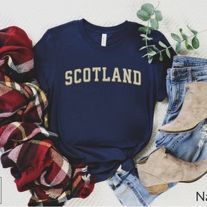 May include: A navy blue t-shirt with the word "SCOTLAND" printed in gold on the front. The shirt is laying on a white surface with a plaid scarf and blue jeans.