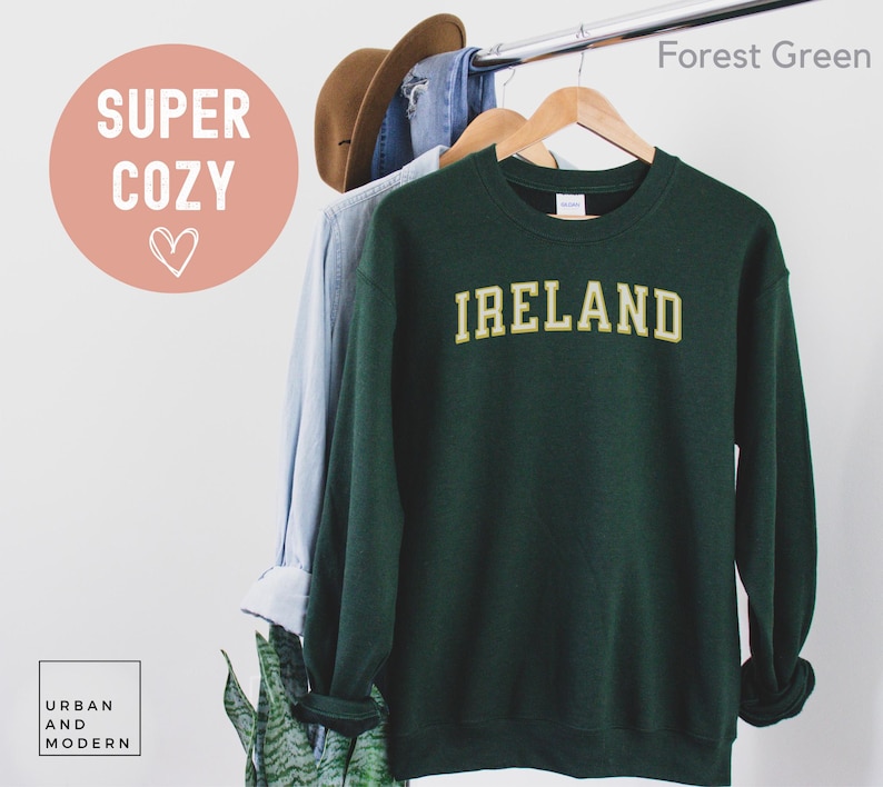 Może przedstawiać: Ciemnozielona bluza z napisem "IRELAND" złotymi literami. Bluza wisi na drewnianym wieszaku z jasnoniebieską koszulą i brązowym kapeluszem. Słowa "SUPER COZY" znajdują się w r&oacute;żowym okręgu.