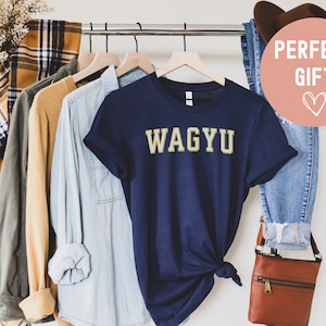May include: Navy blue t-shirt with the word "WAGYU" in gold lettering. The shirt is displayed on a clothing rack with other shirts and a pair of jeans. A brown leather crossbody bag is also visible.
