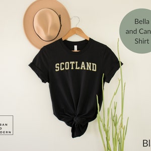 May include: Black t-shirt with the word "SCOTLAND" in gold lettering. The shirt is on a white background with a hat and a plant.