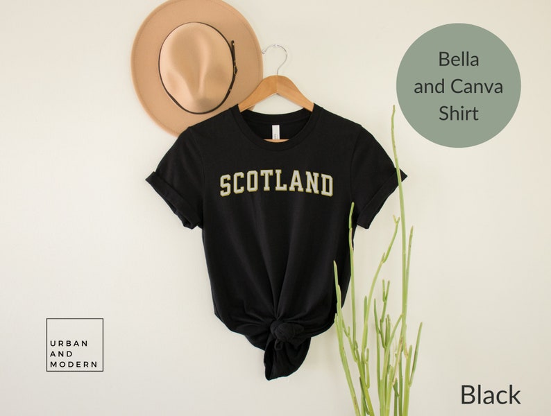 May include: Black t-shirt with the word "SCOTLAND" in gold lettering. The shirt is on a white background with a hat and a plant.