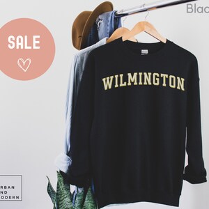 May include: Black sweatshirt with "WILMINGTON" in gold lettering. The word "SALE" is in a pink circle in the upper left corner. A brown hat and blue shirt hang on the rack behind the sweatshirt. The word "Black" is in the upper right corner.