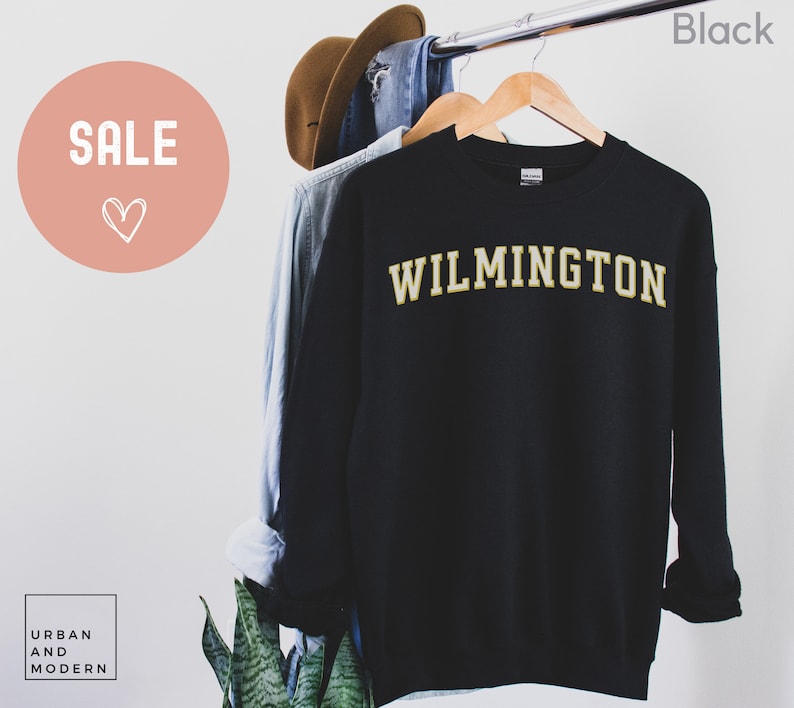 May include: Black sweatshirt with "WILMINGTON" in gold lettering. The word "SALE" is in a pink circle in the upper left corner. A brown hat and blue shirt hang on the rack behind the sweatshirt. The word "Black" is in the upper right corner.