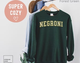 May include: Forest green sweatshirt with the word "NEGRONI" in a gold, collegiate-style font. The sweatshirt is hanging on a wooden hanger with a light blue shirt and a brown hat. The words "SUPER COZY" and a heart are in a pink circle.