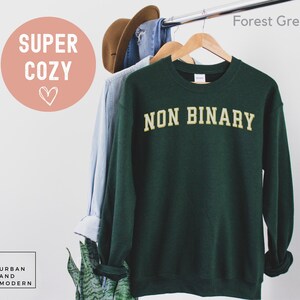 May include: A forest green sweatshirt with the text "NON BINARY" in a gold, collegiate-style font. The sweatshirt is hanging on a wooden hanger. The words "SUPER COZY" are in a pink circle.