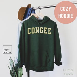 May include: A hunter green hoodie with the word "CONGEE" in a gold and white collegiate font. The hoodie features a front pocket and a drawstring hood. The text "COZY HOODIE" is in a pink circle. The brand logo "URBAN AND MODERN" is in a white square.