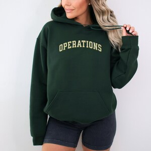 May include: A dark forest green hooded sweatshirt featuring the word OPERATIONS printed in a light beige, arched, collegiate-style font across the chest. The hoodie includes a front kangaroo pocket, long sleeves, and matching green drawstrings.