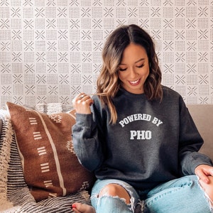 May include: A woman wearing a gray sweatshirt with the text "Powered by Pho" in white lettering. She is sitting on a couch with a brown and white patterned throw pillow.