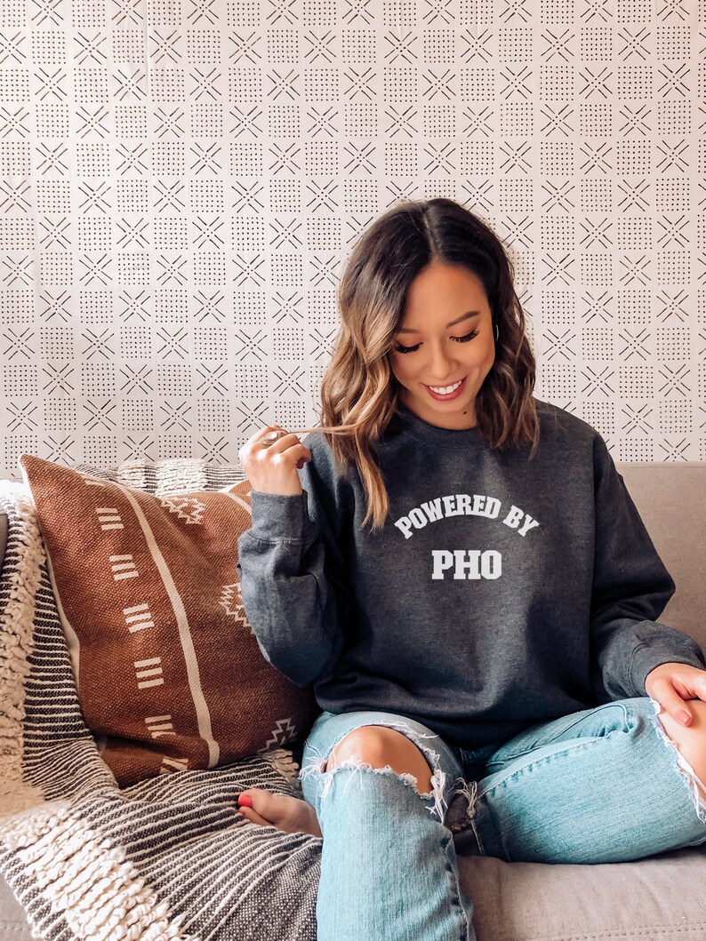 May include: A woman wearing a gray sweatshirt with the text "Powered by Pho" in white lettering. She is sitting on a couch with a brown and white patterned throw pillow.