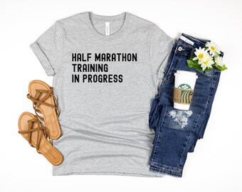 May include: A gray t-shirt with the text "HALF MARATHON TRAINING IN PROGRESS" printed in black. The shirt is paired with blue jeans, a cup of coffee, and brown sandals.