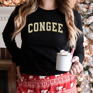 May include: Black long-sleeved sweatshirt with the word "CONGEE" in a yellow and white collegiate font. The person is wearing red and white patterned pyjama bottoms and holding a white mug with marshmallows and a candy cane. A Christmas tree is in the background.