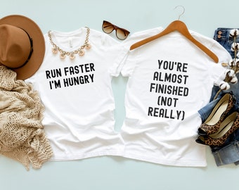 May include: Two white t-shirts with black text. The first shirt says "Run Faster I'm Hungry". The second shirt says "You're Almost Finished (Not Really)".