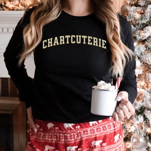 May include: Black long-sleeve shirt with the word "CHARTCUTERIE" in gold lettering. The person is wearing red and white patterned pajama pants and holding a white mug with marshmallows and a candy cane. A Christmas tree is in the background.
