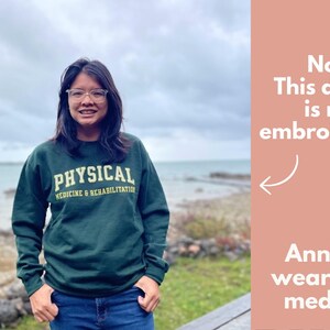 May include: A dark green sweatshirt with the text "PHYSICAL MEDICINE & REHABILITATION" in gold lettering. The sweatshirt is worn by a person wearing glasses and blue jeans. The background shows a body of water and a cloudy sky.