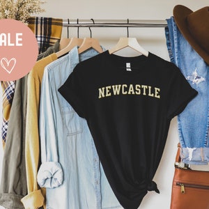 May include: A black t-shirt with the word "NEWCASTLE" in gold lettering. The shirt is displayed on a hanger with other clothing items, including a denim shirt, jeans, and a brown hat. A "SALE" graphic is visible.