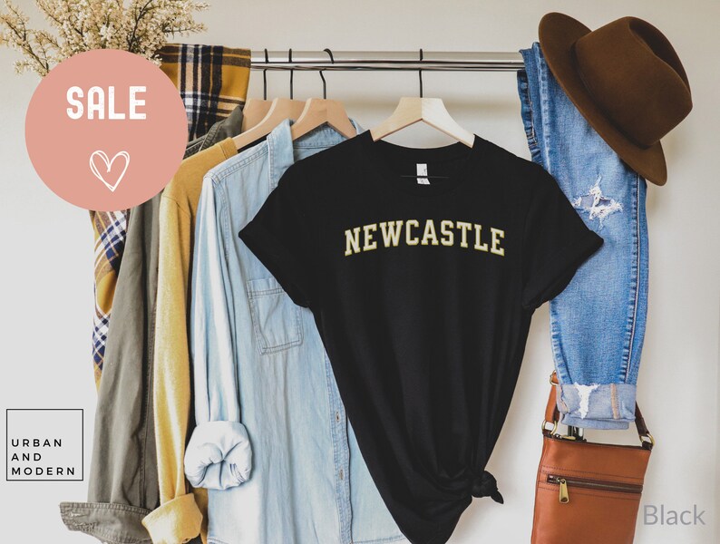 May include: A black t-shirt with the word "NEWCASTLE" in gold lettering. The shirt is displayed on a hanger with other clothing items, including a denim shirt, jeans, and a brown hat. A "SALE" graphic is visible.