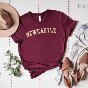 May include: Maroon t-shirt with "NEWCASTLE" in gold lettering. The shirt is styled with a beige hat, light-wash jeans, and brown flats. A string of wooden beads and greenery complete the casual outfit. The Urban and Modern logo is in the bottom left corner.