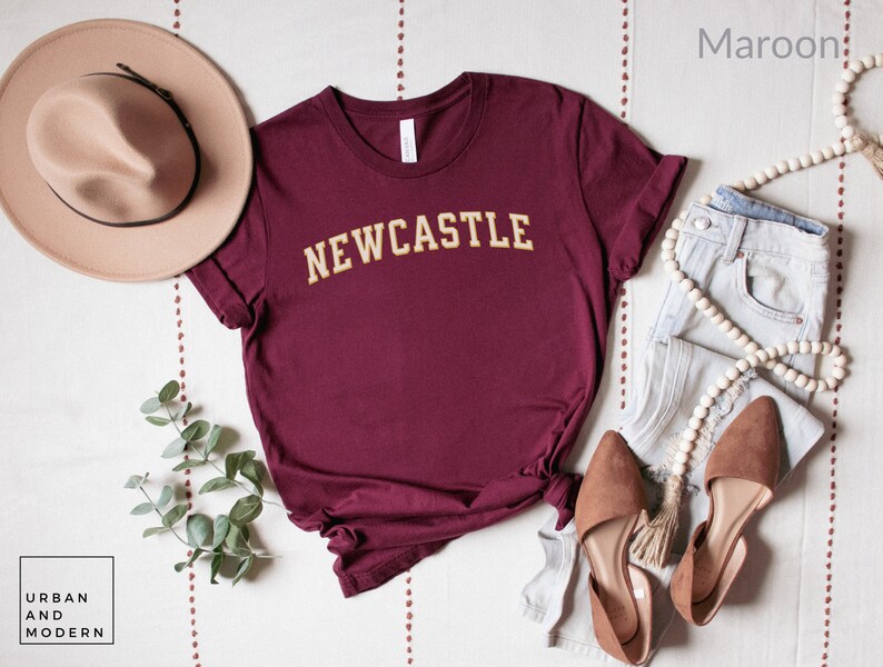 May include: Maroon t-shirt with "NEWCASTLE" in gold lettering. The shirt is styled with a beige hat, light-wash jeans, and brown flats. A string of wooden beads and greenery complete the casual outfit. The Urban and Modern logo is in the bottom left corner.