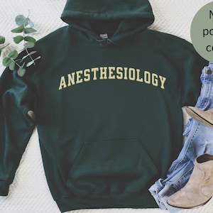 May include: A dark green hooded sweatshirt with the text "ANESTHESIOLOGY" in a gold color on the front. The sweatshirt is laid flat on a white surface with a pair of blue jeans and brown ankle boots.