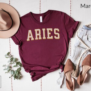 Op de afbeelding: Bordeauxrood t-shirt met het woord "ARIES" in gouden en cr&egrave;mekleurige blokletters. Het shirt is gestyled met een beige hoed, lichtblauwe jeans en bruine ballerina's. De afbeelding bevat ook een snoer van houten kralen en een takje groen.