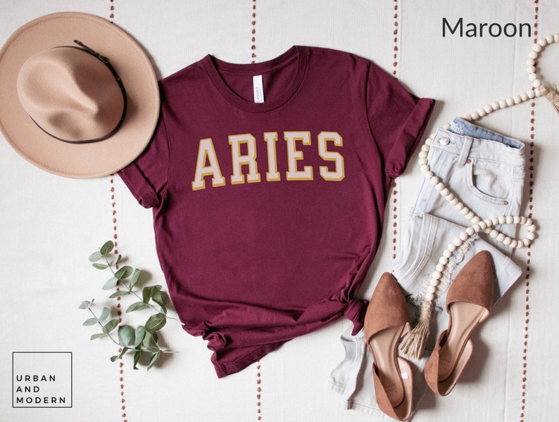 Op de afbeelding: Bordeauxrood t-shirt met het woord "ARIES" in gouden en cr&egrave;mekleurige blokletters. Het shirt is gestyled met een beige hoed, lichtblauwe jeans en bruine ballerina's. De afbeelding bevat ook een snoer van houten kralen en een takje groen.
