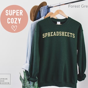 May include: A forest green sweatshirt with the word "SPREADSHEETS" in a gold, collegiate-style font. The sweatshirt is hanging on a wooden hanger with other clothing items. The words "SUPER COZY" are in a pink circle.