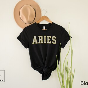 Op de afbeelding: Zwart T-shirt met het woord "ARIES" in een gouden en witte college-lettertype. Het shirt wordt gepresenteerd op een houten hanger met een beige hoed op de achtergrond. De woorden "Urban and Modern" staan in een witte doos linksonder.